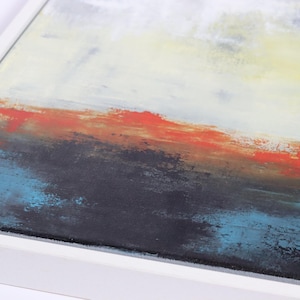May include: Abstract landscape painting in a white frame. The artwork features a layered composition with a color palette of white, yellow, orange, black, and blue. The brushstrokes create a textured effect, evoking a sense of depth and movement.