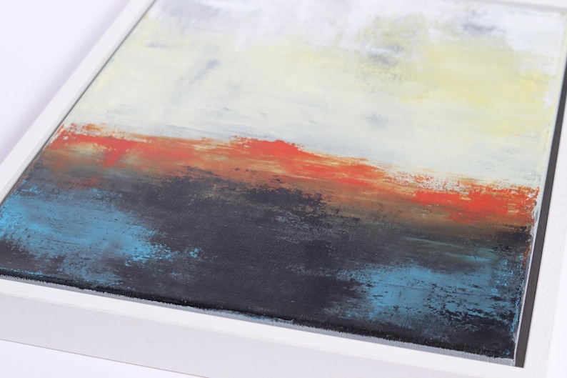 May include: Abstract landscape painting in a white frame. The artwork features a layered composition with a color palette of white, yellow, orange, black, and blue. The brushstrokes create a textured effect, evoking a sense of depth and movement.