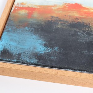 May include: Abstract art piece with a wooden frame. The artwork features a textured surface with layers of color, including orange, blue, black, and white. The painting has a modern aesthetic.