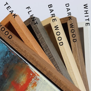 May include: Close-up of a selection of wooden picture frames in various colors, including light wood, teak, flint, bare wood, dark wood, white, and black. The frames are angled, showcasing the different wood tones and textures. The text on the frames indicates the color.