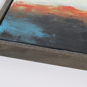 May include: Abstract art piece with a dark gray wooden frame. The canvas features a textured surface with a blend of colors, including black, blue, orange, and white. The artwork is a close-up view, showcasing the details of the paint.