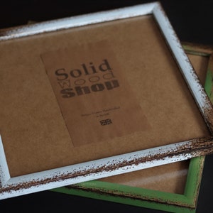 May include: Two rustic picture frames, one with a white distressed finish and the other with a green distressed finish. The white frame has a brown paper backing with a label that reads "Solid Wood Shop, Picture Frames Handcrafted in the UK".