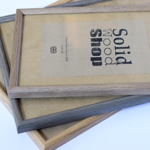 Puede incluir: Dos marcos de fotos de madera apilados uno encima del otro. El marco superior es de madera marr&oacute;n claro con un paspart&uacute; blanco y el texto "Solid Wood Shop" impreso en el paspart&uacute;. El marco inferior es de madera gris oscuro con un paspart&uacute; blanco.