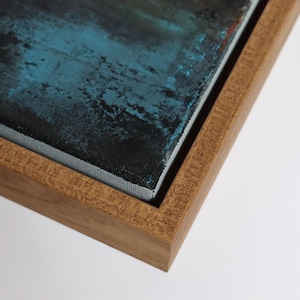 May include: Close-up of a framed abstract painting. The artwork features a textured surface with shades of blue and black. The painting is encased in a natural wood frame, showcasing the corner and edges of the piece.
