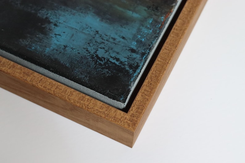 May include: Close-up of a framed abstract painting. The artwork features a textured surface with shades of blue and black. The painting is encased in a natural wood frame, showcasing the corner and edges of the piece.