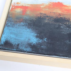 May include: Abstract painting with a textured surface. The artwork features a blend of colors, including orange, blue, black, and white, within a natural wood frame. The brushstrokes create a sense of depth and movement.