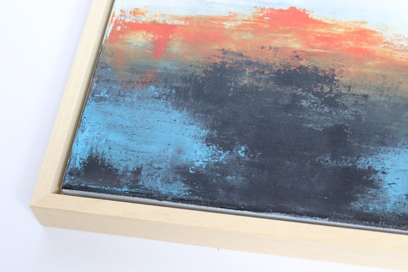 May include: Abstract painting with a textured surface. The artwork features a blend of colors, including orange, blue, black, and white, within a natural wood frame. The brushstrokes create a sense of depth and movement.