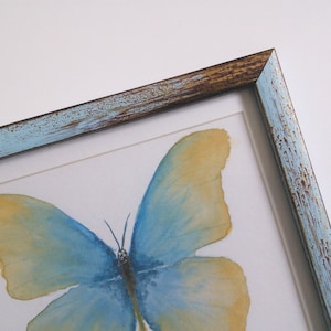 May include: A framed watercolor painting of a blue and yellow butterfly with a distressed blue and brown frame.