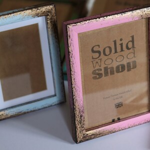 May include: Two wooden picture frames with distressed paint. The frame on the left is light blue with a white mat. The frame on the right is pink with a brown mat. The frame on the right has a brown paper insert with the text "Solid Wood Shop" printed on it.