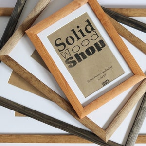 May include: Four wooden picture frames stacked on top of each other. The top frame is light brown and has the text "Solid Wood Shop" and "Picture Frames Handcrafted in the UK" printed on a brown paper insert. The frames below are a darker brown, grey, and a lighter brown.