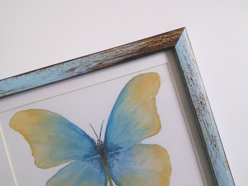 May include: A framed watercolor painting of a blue and yellow butterfly with a distressed blue and brown frame.