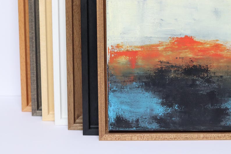 May include: Abstract art piece with a textured surface, featuring a color palette of orange, black, and blue. The artwork is displayed in a natural wood frame, with a variety of other frame colors visible in the background.