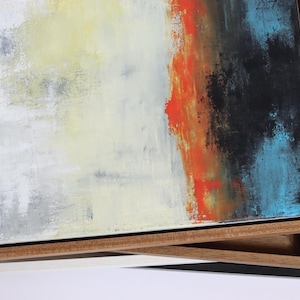 May include: Abstract art canvas with a wooden frame. The artwork features a blend of colors, including white, yellow, orange, black, and blue. The canvas is stacked on top of other framed canvases with different colored frames.