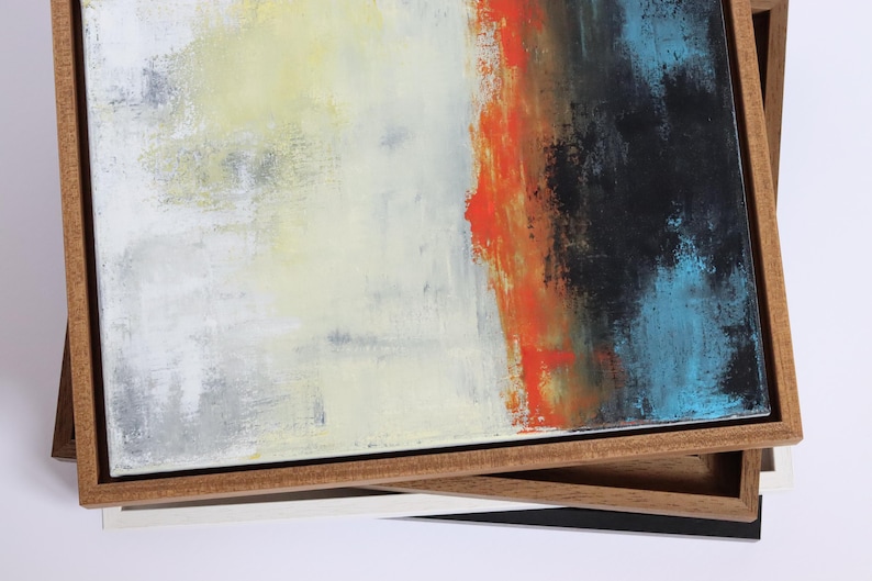 May include: Abstract art canvas with a wooden frame. The artwork features a blend of colors, including white, yellow, orange, black, and blue. The canvas is stacked on top of other framed canvases with different colored frames.