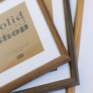 May include: Three wooden picture frames stacked on top of each other. The top frame is light brown, the middle frame is dark brown, and the bottom frame is a lighter brown. The top frame has a white mat and a brown paper insert with the text "solid wood shop" and "Picture Frames Handcrafted in the UK".