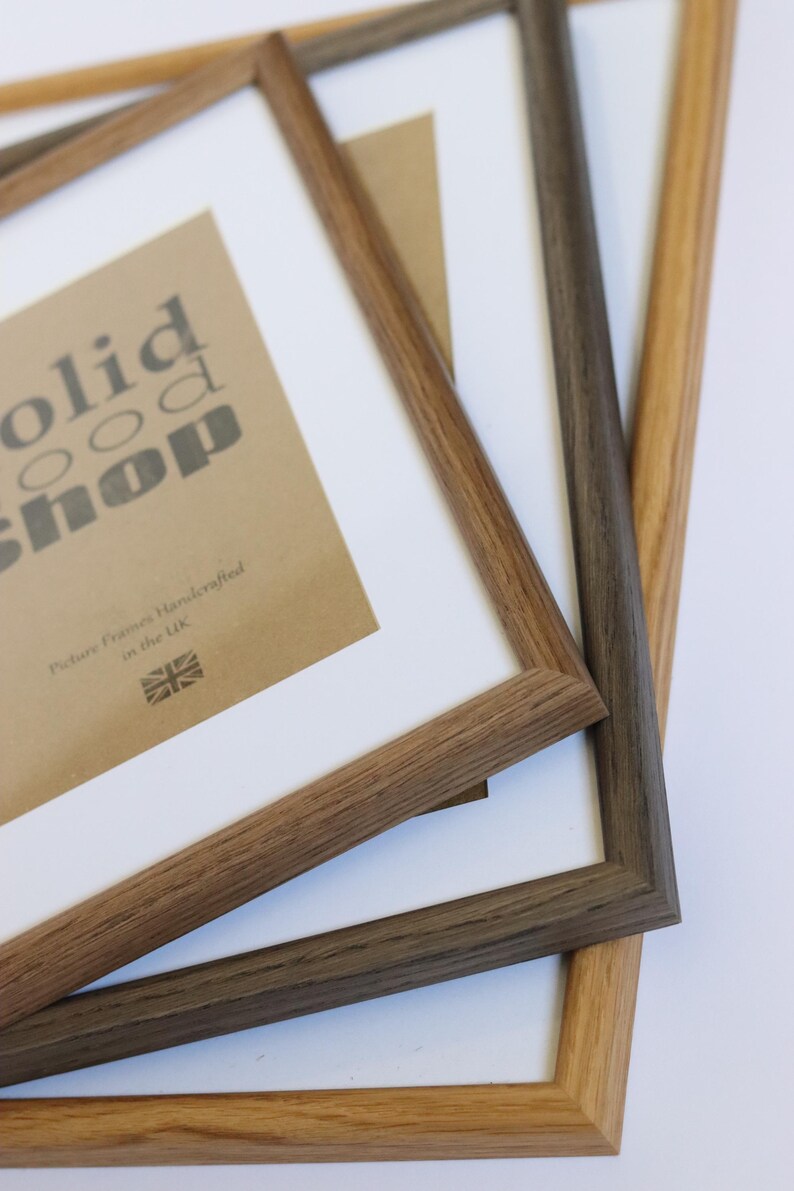 May include: Three wooden picture frames stacked on top of each other. The top frame is light brown, the middle frame is dark brown, and the bottom frame is a lighter brown. The top frame has a white mat and a brown paper insert with the text "solid wood shop" and "Picture Frames Handcrafted in the UK".