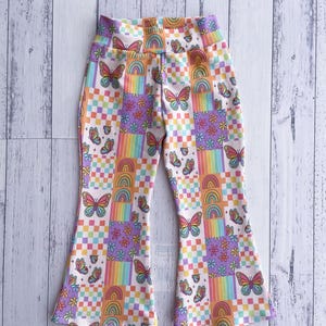 May include: Colorful bell-bottom pants with a repeating pattern of butterflies, rainbows, and flowers. The design incorporates a checkerboard pattern in various pastel colors. The pants have a high waist and flared legs.