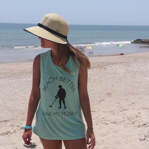 May include: A woman wearing a light blue tank top with a black graphic of a person holding a metal detector. The graphic says "Beach Better Have My Money". She is wearing a wide-brimmed straw hat and is standing on a sandy beach.