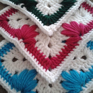 Puede incluir: Tres cuadrados de ganchillo con bordes de diferentes colores. Los cuadrados son blancos con un diseño de flores verdes, rojas y azules en el centro.