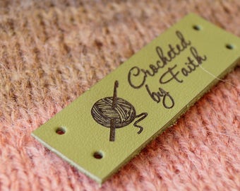 May include: A green leather label with the text "Crocheted by Faith" and a small illustration of a ball of yarn and a crochet hook.