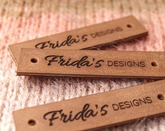 Może przedstawiać: Trzy brązowe skórzane etykiety z wygrawerowanym czarnym tekstem "Frida's Designs".