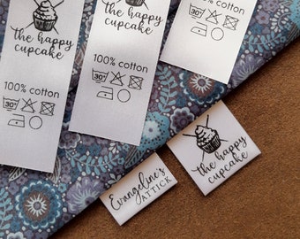 Könnte beinhalten: Vier weiße Stoff-Etiketten mit schwarzem Text und einer Cupcake-Grafik. Die Etiketten lauten "the happy cupcake" und "100% Baumwolle". Die Etiketten enthalten auch Pflegehinweise zum Waschen, Trocknen und Bügeln. Die Etiketten befinden sich auf einem blau-weißen Blumenstoff.