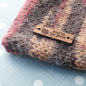 May include: A brown, beige, and pink striped knitted hat with a wooden tag that reads "knits by jane" and a small image of yarn and knitting needles.