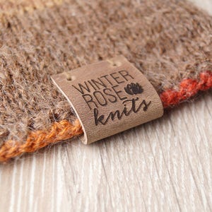 Può includere: Un'etichetta in pelle marrone con il testo "Winter Rose Knits" in lettere nere. L'etichetta è attaccata a un tessuto a maglia marrone e arancione.