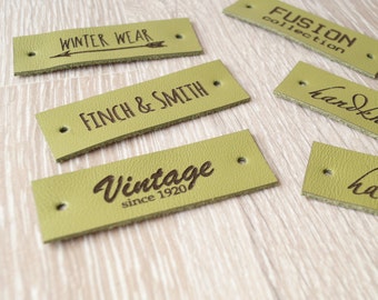 May include: Five green leather labels with different text. The labels are on a light wood surface. The text on the labels reads: "Winter Wear", "Finch & Smith", "Vintage since 1920", "Fusion Collection", and "Handmade".