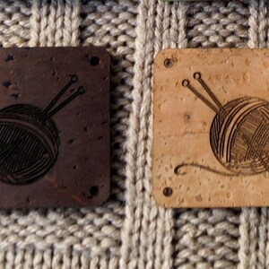 May include: Two square cork coasters with a laser-etched design of a ball of yarn and knitting needles. The coasters are shown in two colors: dark cork and natural cork.