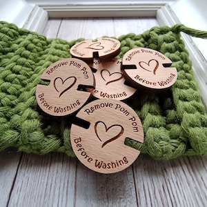 May include: Several wooden circular tags with the text "Remove Pom Pom Before Washing" and a heart design. The tags are light brown and are placed on a green crocheted fabric. These tags are for a craft project.