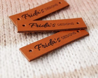 May include: Three brown leather labels with the text "Frida's Designs" embossed in black.