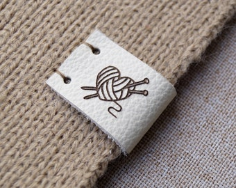 May include: A white leather tag with a black outline of a ball of yarn and knitting needles. The tag is attached to a brown knitted fabric.