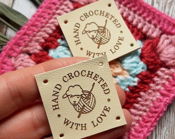May include: Two beige leather labels with holes for stitching. Each label has a brown circle with the text "Hand crocheted with love" and a small illustration of a sheep holding yarn.