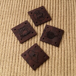 May include: Four brown cork fabric patches with different designs. The patches have holes in the corners. The designs are an owl, a ball of yarn with knitting needles, a heart shape, and the words "Be cool" in a circle.