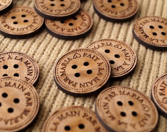 May include: Wooden buttons with the text "La Main Par Maria Fait A La Main" in brown lettering. The buttons are round and have a natural wood finish.