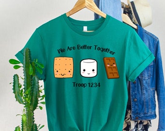 May include: A green t-shirt featuring a graphic of a graham cracker, marshmallow, and chocolate bar, representing a s'more. The text "We Are Better Together" and "Troop 1234" is printed on the shirt.