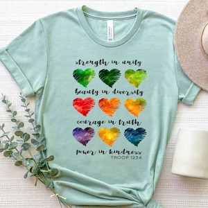 May include: A light blue t-shirt with a rainbow heart design and the text "strength in unity", "beauty in diversity", "courage in truth", "power in kindness", and "TROOP 1234".