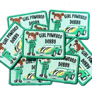 May include: A pile of green and black embroidered iron-on patches with the text "Girl Powered Derby" and a cartoon girl in a racing suit driving a green race car.