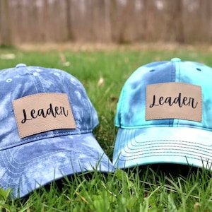 May include: Two tie-dye baseball caps with brown leather patches that say "Leader" on them. The caps are on a bed of green grass.
