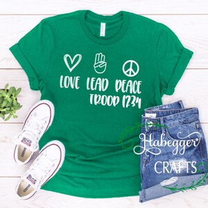 May include: Green t-shirt with white text and graphics. The text reads "Love Lead Peace Troop 1234". The graphics are a heart, a peace sign, and a hand with two fingers raised.