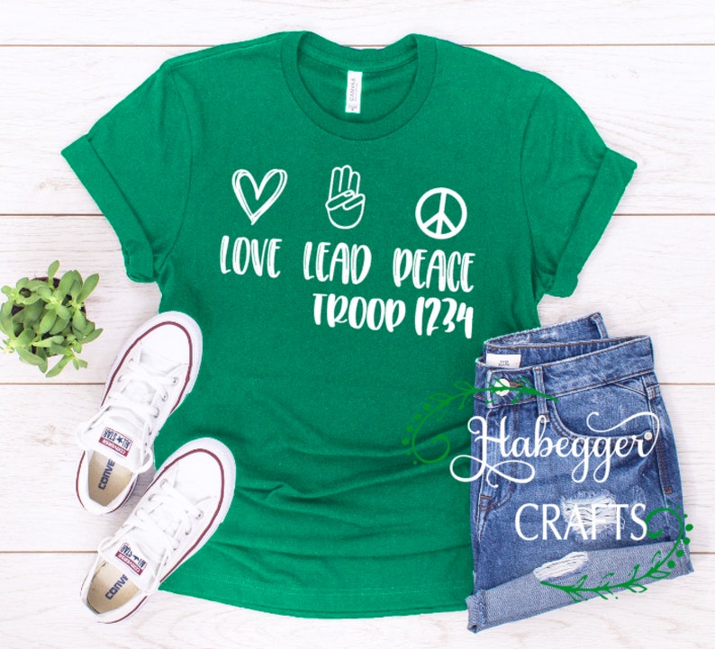 May include: Green t-shirt with white text and graphics. The text reads "Love Lead Peace Troop 1234". The graphics are a heart, a peace sign, and a hand with two fingers raised.