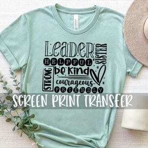 May include: A light blue t-shirt with a black screen print design that says "Leader", "Helpful", "Be Kind", "Strong", "Courageous", "Friendly", and "Sister". The text is arranged in a square shape with a heart in the center. The shirt is on a white surface with a white mug and a straw hat.