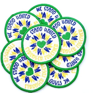 May include: A stack of eight round, white embroidered patches with green borders. Each patch features a yellow and blue sunflower design with a green heart in the center. The text "We Stand United" and "Stronger Together" is embroidered in blue and yellow around the sunflower design.