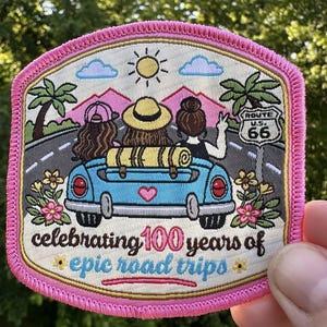 May include: Embroidered patch with a pink border featuring a blue car carrying three people on a road trip. The patch includes palm trees, mountains, a sun, and the text "Route 66" and "celebrating 100 years of epic road trips."