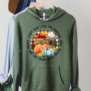 May include: Green hooded sweatshirt with a white and black checkered background with a graphic of pumpkins, apples, and a coffee cup. The text reads "Warm Coffee and Cool Nights Every Year I Fall for Pumpkin Apple Picking".