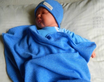 May include: A baby doll wearing a blue knitted hat and wrapped in a matching blue blanket. The blanket has a tag that reads "Anna Maria". The doll is lying on a bed with a light-coloured pillow.