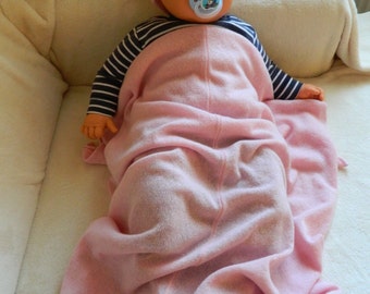 May include: A pink baby blanket with a white label that says "Lambswool". A baby doll wearing a pink hat and a dummy is lying on the blanket.