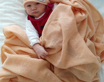 May include: A baby wearing a peach-coloured knitted hat with the text "Anna Maria" on it, is wrapped in a peach-coloured knitted blanket with the same text on it. The baby is lying on a white surface.