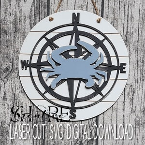 May include: A black and white compass with a blue crab silhouette in the center. The compass has the directions North, South, East, and West. The background is a white wooden plank.